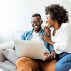 A couple laughs together as they sit on a couch and use a laptop.